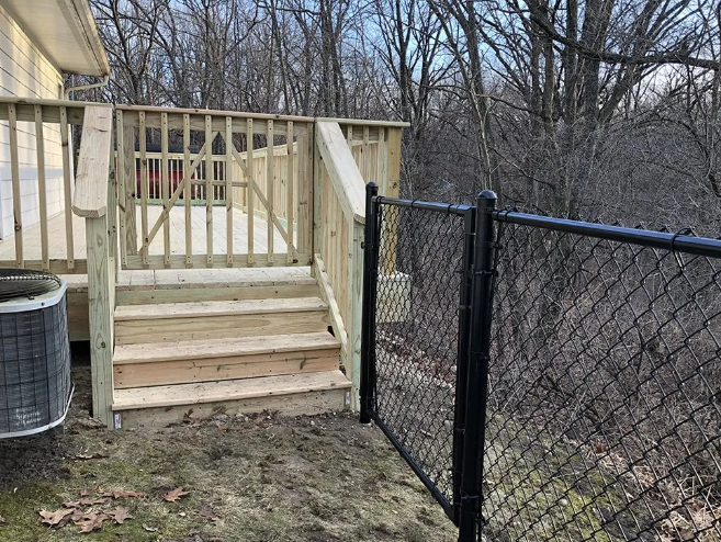 Wooden deck with stairs, chain-link fence, and air conditioning unit against a wooded backdrop.