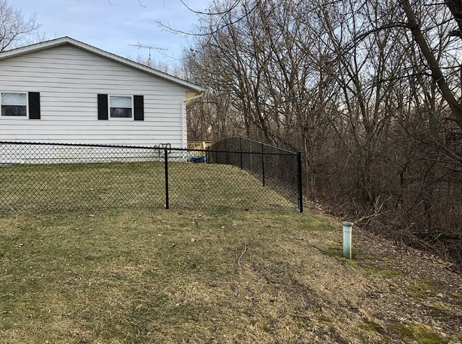 Black chain-link fence bordering a grassy yard next to a white house and wooded area.