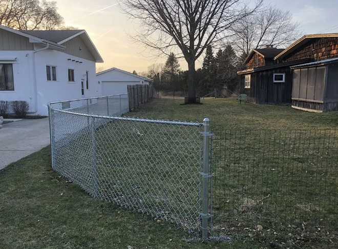 Chain-link fence curves in a grassy yard, between two buildings, under a cloudy sky.