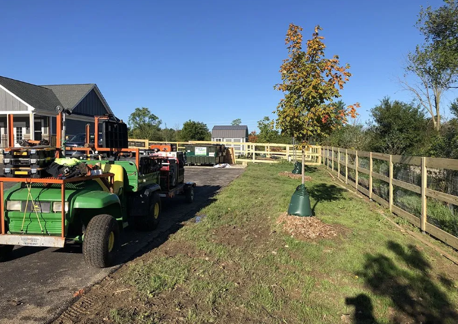 Green utility vehicle parked next to a new wooden fence, grass, and a residential area on a sunny day.