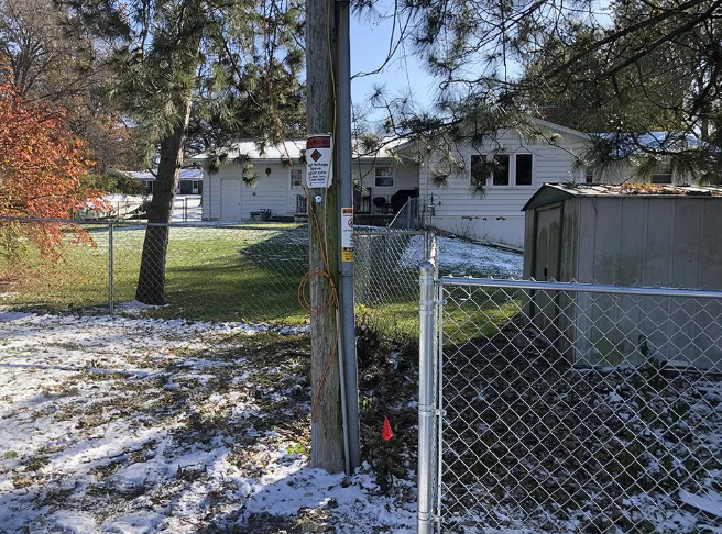 Snowy backyard with fence, power pole with camera, and a white house in the distance.