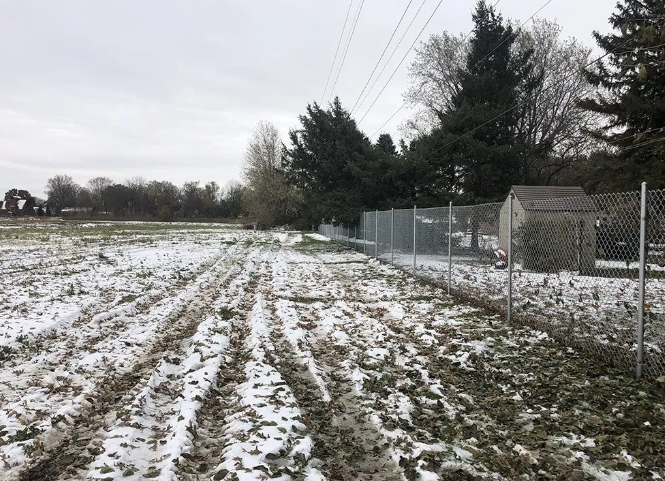 Snow-covered field with tire tracks, a chain-link fence, and a shed. Power lines and trees are in the background.