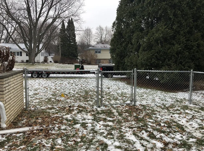 Snow-covered backyard with a chain-link fence. A trailer is parked in the distance. Overcast sky.