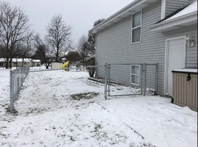 Snowy backyard with a chain-link fence, gate, and the side of a two-story house.
