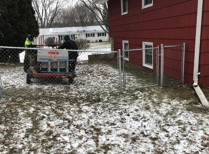 Two people moving a machine near a chain-link fence by a red building, snow on the ground.