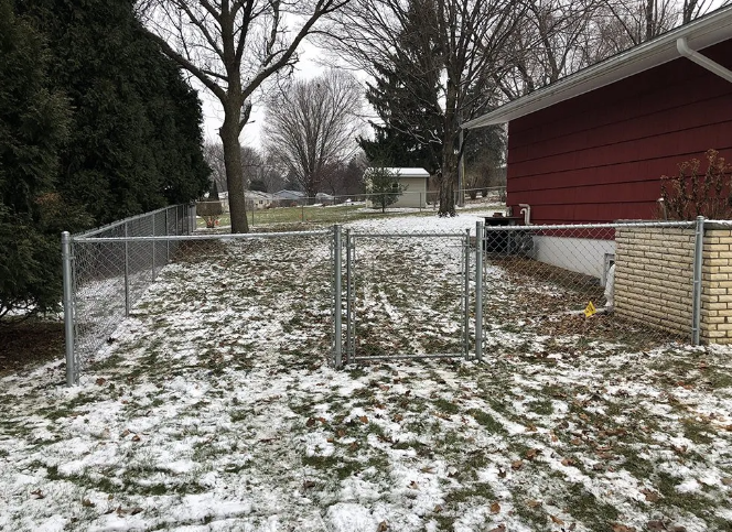 Chain link fence in a snow-covered yard, with a gate, next to a red-sided building.