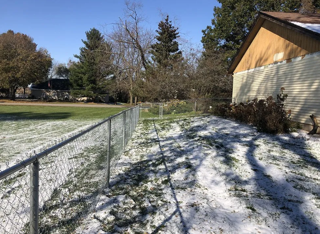 Snowy backyard with a chain-link fence, a house, and some trees.