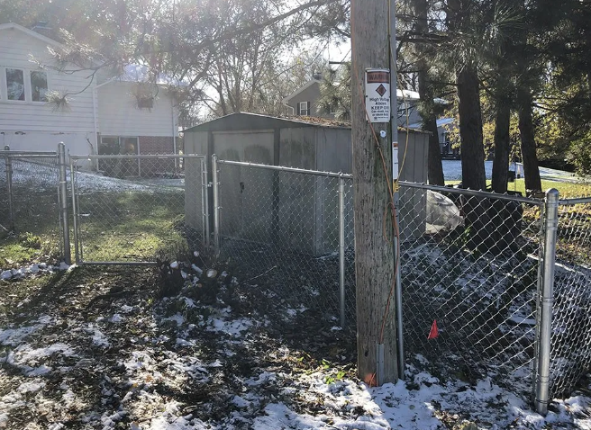 Chain-link fence surrounds a metal shed, partially covered in snow, next to a utility pole, and a small house.