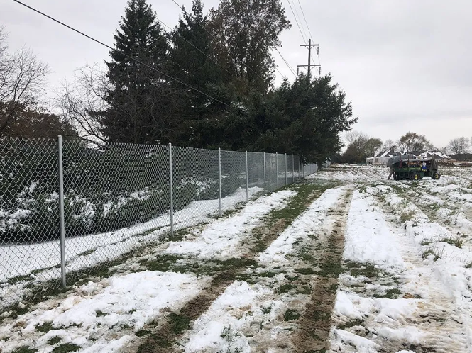 Chain-link fence borders a snowy dirt road. Trees and power lines are visible on a cloudy day.