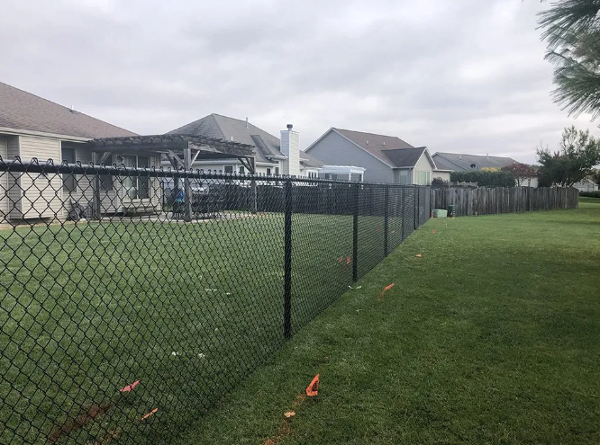 Black chain-link fence in a grassy backyard, adjacent to several houses under an overcast sky.