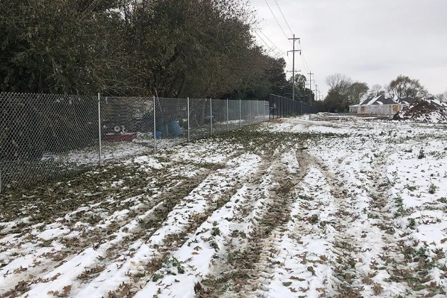 Snowy field with muddy tire tracks, chain-link fence, and trees under overcast sky.