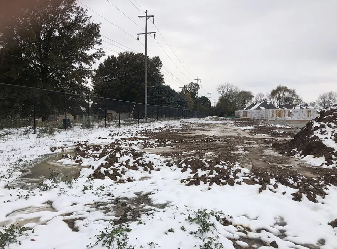 Snowy, muddy construction site with dirt piles and power lines; fence and houses in background.