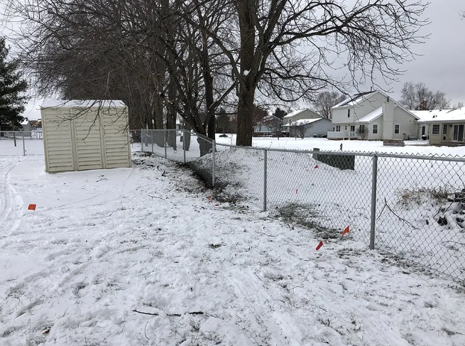 Snowy backyard with chain-link fence, beige shed, and houses in the background.