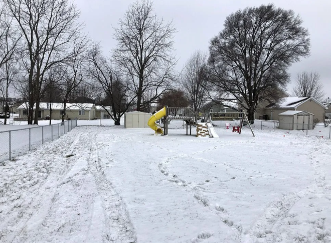 Snow-covered playground with a yellow slide and swings, surrounded by a fence and bare trees under a cloudy sky.