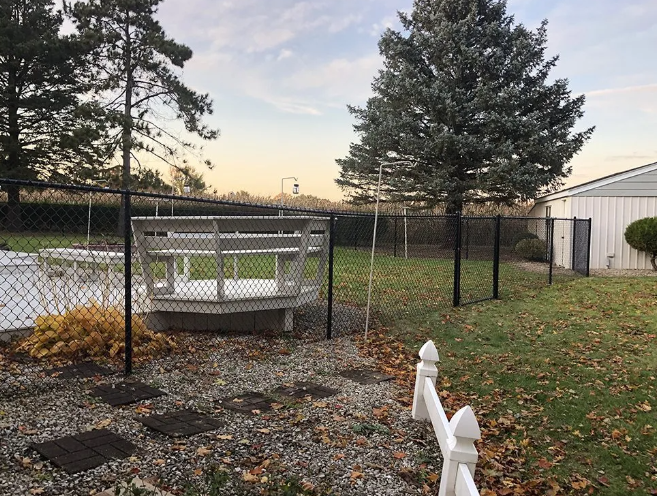 Black chain-link fence surrounds a backyard with a deck, trees, and stone path under a cloudy sky.