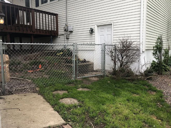 Chain-link fence with gate beside a house. Green grass and stepping stones in front. Deck and white door in the background.