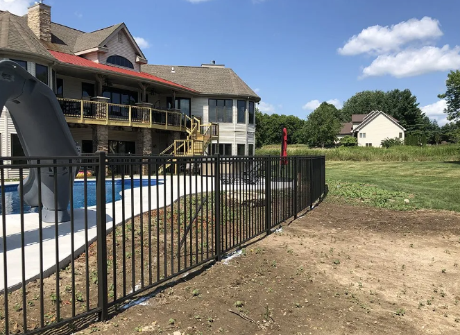 Black fence encloses a pool area next to a large house, a yard in the background.