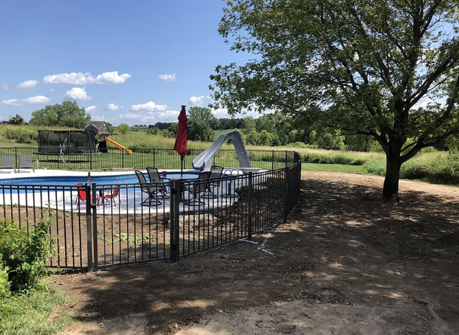 Black fenced-in pool with a slide, red umbrella, and chairs on a sunny day.