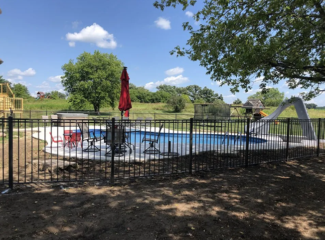 Swimming pool surrounded by a black fence and concrete, with a red umbrella, chairs, and slide.