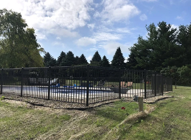 Black fenced outdoor swimming pool on grassy land; trees and cloudy sky in background.