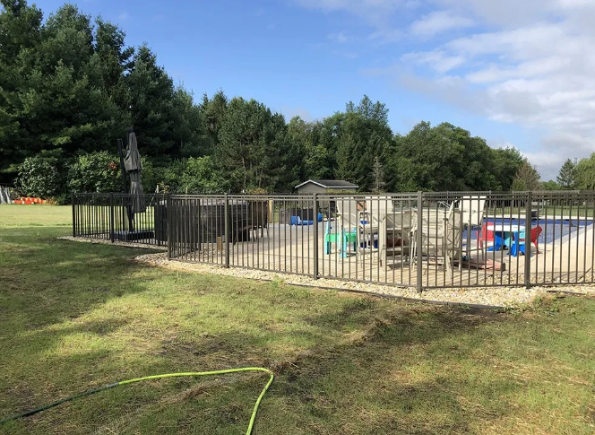 A fenced-in backyard pool area with trees in the background under a blue sky.