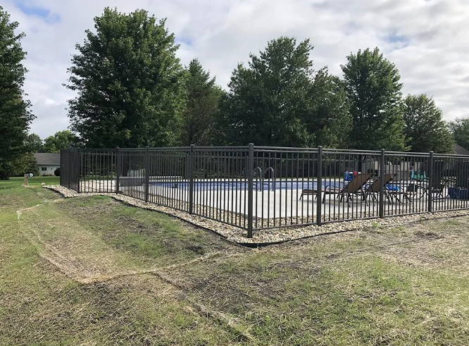 A fenced-in above-ground pool with surrounding landscaping. Brown metal fence, trees in background, cloudy sky.