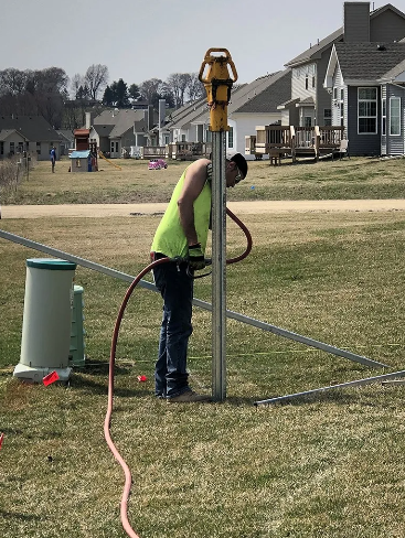 Construction worker using a tool on a metal pole in a grassy yard. Houses in the background.