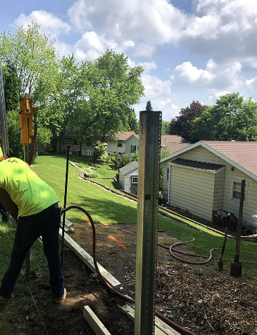 Construction worker installs fence post near a yard with houses and green trees under a cloudy sky.