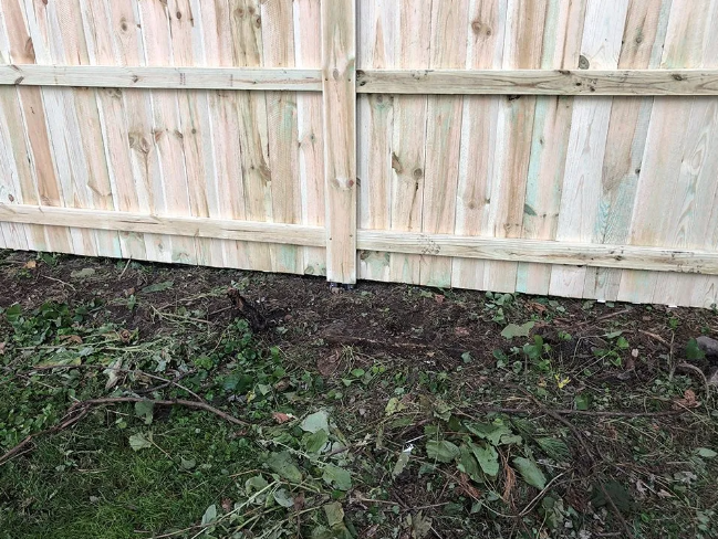Wooden fence, light brown, next to dark soil and weeds.