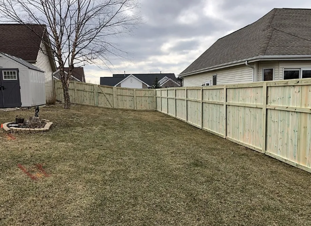 A backyard with a wooden fence surrounding a grassy area, a shed, and houses. Cloudy sky.