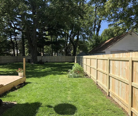 Lawn with a wooden fence around it. A small deck is in the foreground. Trees and a house in the background.