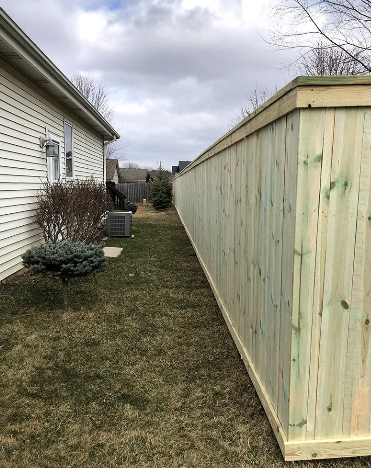 A wooden privacy fence extends alongside a house on a grassy lawn under a cloudy sky.