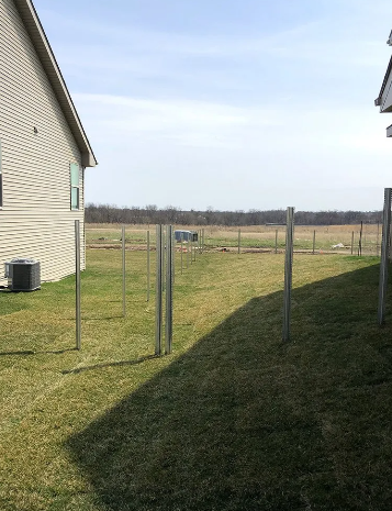 Metal fence posts in a grassy yard, with a house on the left and field in the distance.