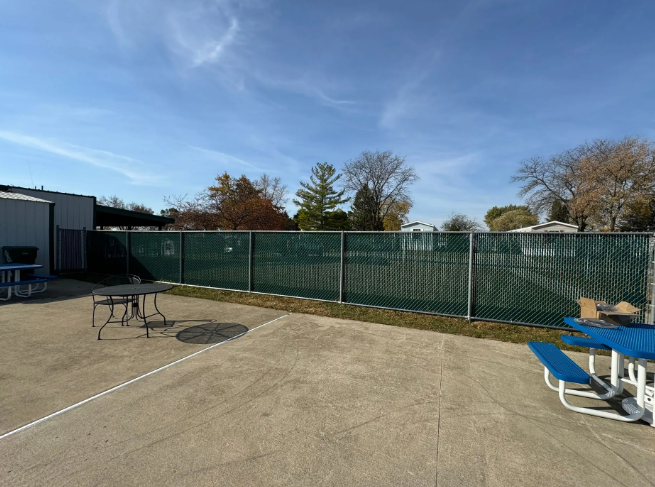 A green privacy fence surrounds a concrete patio with picnic tables, under a blue sky.