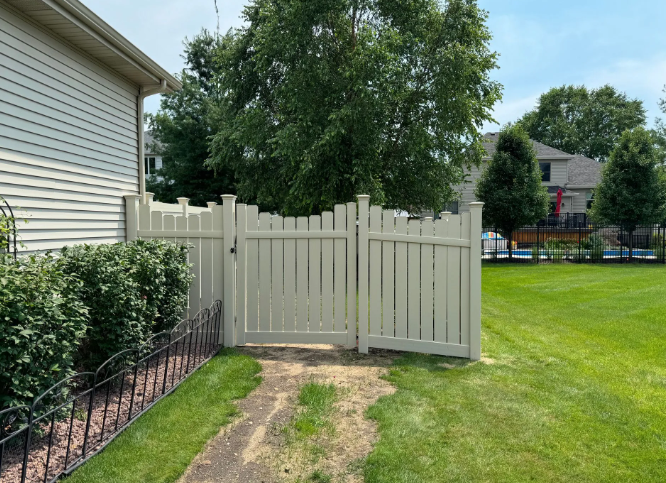 Beige picket fence with gate, leading to a backyard with green grass and trees.