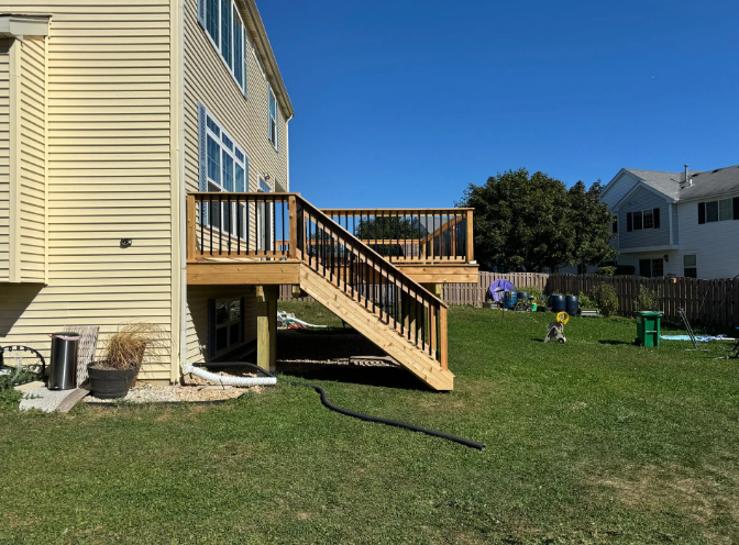 A two-story house with a wooden deck and steps in a grassy backyard under a blue sky.