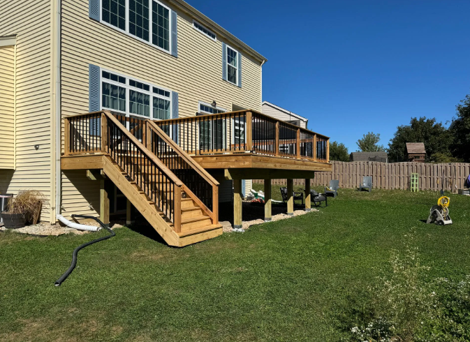 Wooden deck attached to a two-story beige house with black railing, stairs to the yard, and green grass.