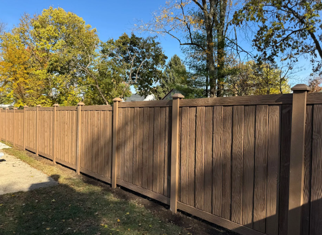 Brown wood-like privacy fence in yard, sunny day, with trees in background.