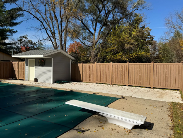 Swimming pool with green cover, diving board, tan fence, and small shed on a sunny day.