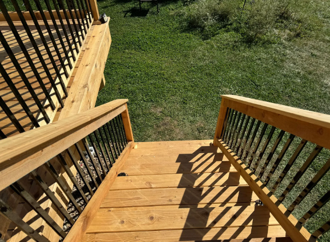 Wooden deck stairs leading to a grassy yard, with black railing spindles and wood handrails.
