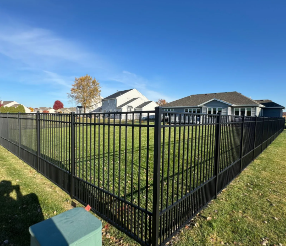 Black metal fence encloses a green lawn with houses under a blue sky.