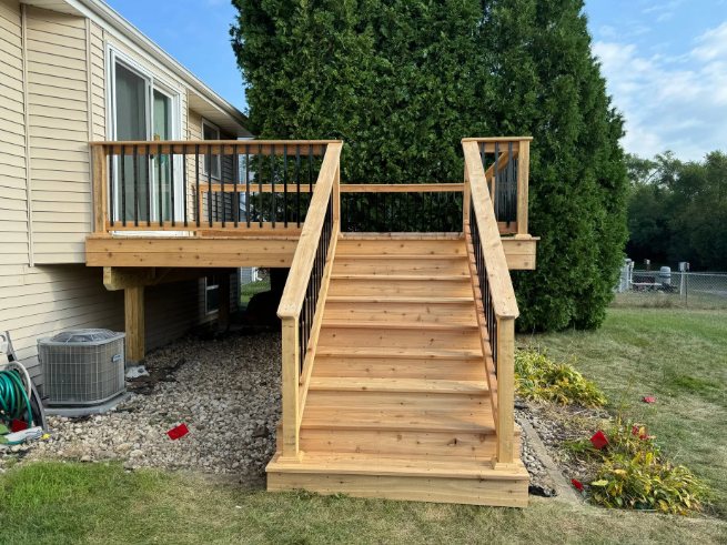 Wooden deck with stairs leading down to a yard; brown and black railings; next to a house with an air conditioner.