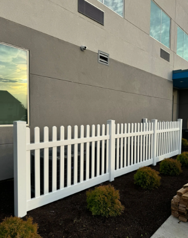 White picket fence along a gray building with bushes and dark mulch.