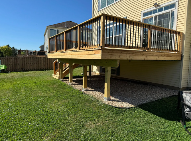 Wooden deck attached to a yellow house, with steps leading down to a gravel area on a grassy lawn.