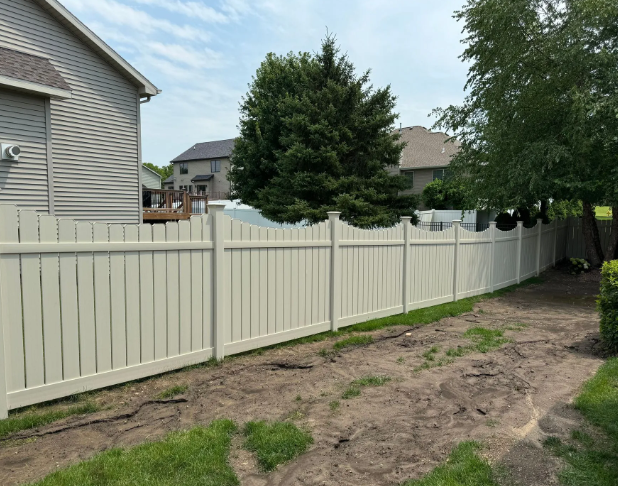 A beige vinyl fence separates two yards, under a cloudy sky.