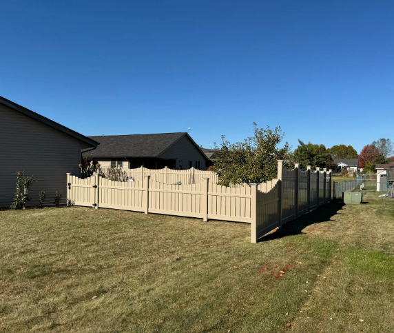 Tan vinyl fence in a backyard, partially enclosing a grassy area near a house and trees under a blue sky.