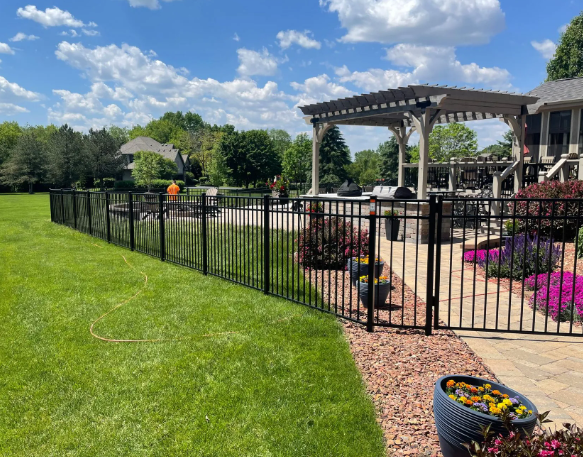 Black metal fence surrounds a lush green lawn. A patio with a pergola and landscaping is visible beyond the fence.