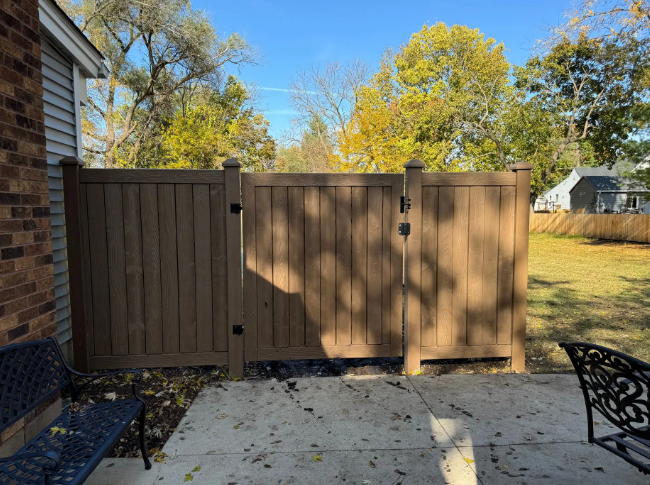 Brown privacy fence with gate on a concrete patio, with a brick building and foliage in the background.