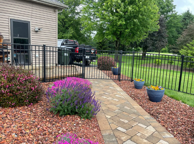 Black metal fence surrounding a yard with a brick pathway, purple flowers, and blue planters. A black truck is visible.