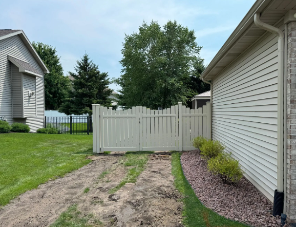 A white vinyl fence with a gate in a residential backyard. Dirt path leads to the gate.